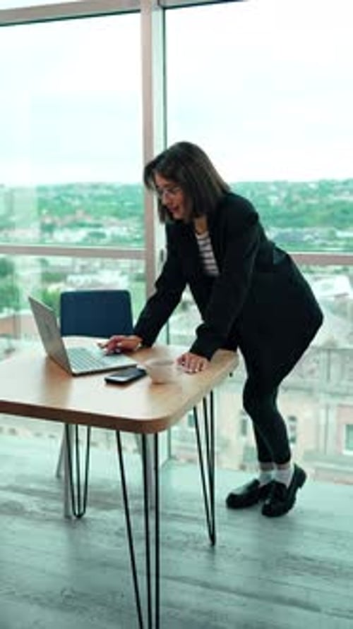 Modern woman stands leaning on the desk using her laptop. Business lady working in the office.