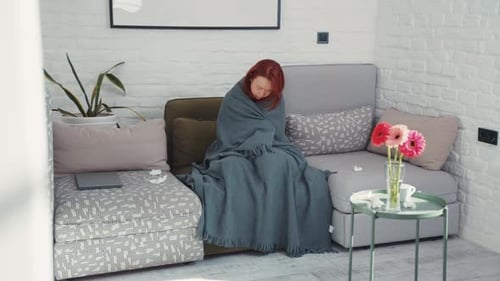 Jeune femme malade asiatique frissonnant de froid et se réchauffant sous une couverture dans un salon confortable