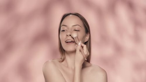 Joyful Seminude Woman Smiling Uses a Makeup Brush in the Studio on a Pink Background A Woman Does