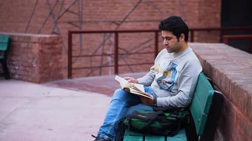 Male Bookworm Enjoying Nature While Reading, Young South Asian Man Engrossed in Reading Outdoors.