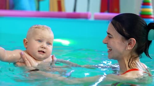 Brunette lady is holding a blond baby in the swimming pool.