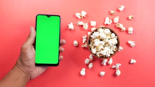 Holding Smart Phone with Green Screen and Popcorn on Red Background
