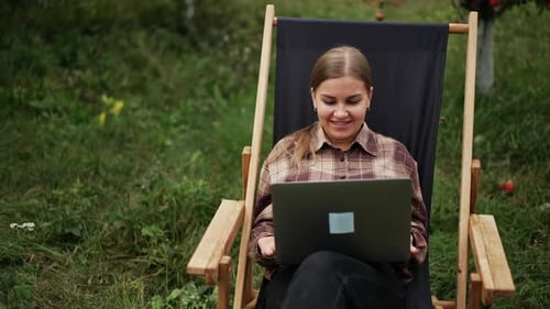 Positiv lächelnde junge Frau sitzt im Gartenstuhl und arbeitet am Laptop.