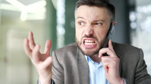 Angry dissatisfied businessman arguing talking on phone sitting at workplace in business office.