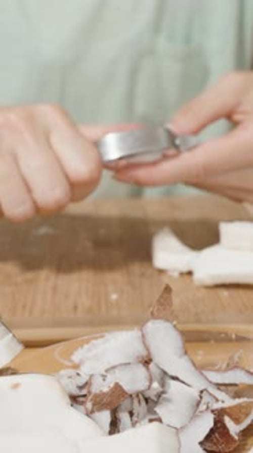 Vertical video. Using a Vegetable Peeler, the Woman Removes the Brown Skin from the Coconut Flesh.