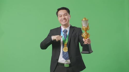 Asiatischer Geschäftsmann zeigt Goldtrophäe vor die Kamera, lächelt und zeigt auf sich selbst auf den grünen Bildschirm