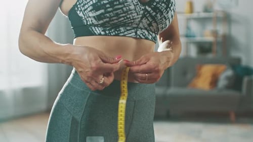 Strong and Beautiful Fit Girl in an Athletic Top is Measuring Her Waist With a Tape Ruler in Her Br