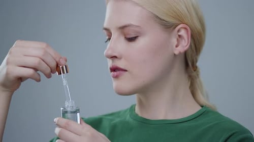 Attractive Blonde Girl Smiling Applying Fake Tears on Her Nude Face on Grey Background Portrait of