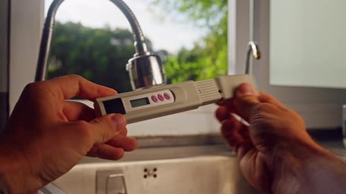 First Person View Of Hand Holding Digital Water Quality Tester Near Kitchen Faucet With Tropical