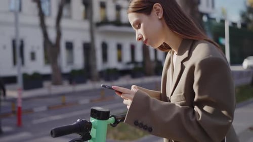 Young Woman Using Smartphone Next to an Electric Scooter in the City
