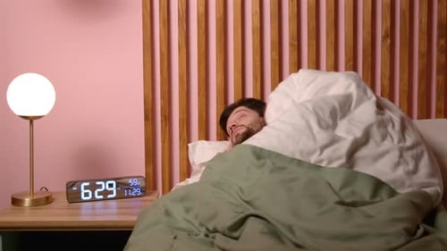 Ausgeruhter positiver Mann mittleren Alters, der morgens mit Wecker im Bett aufwacht