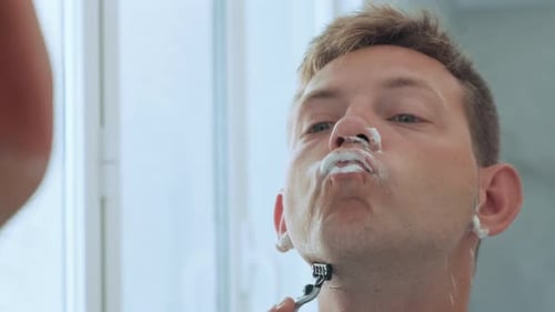 Caucasian Handsome Young Man Shaving His Beard Standing in the Bathroom and Looking at Mirror Daily