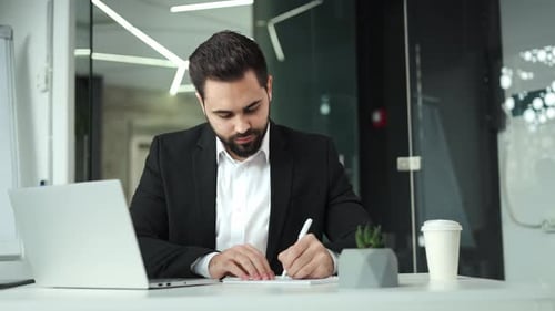 Entrepreneur Typing on Laptop and Noting Information on Piece of Paper in Office