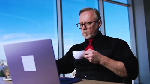 Calm confident bearded man works on laptop and drinks coffee.