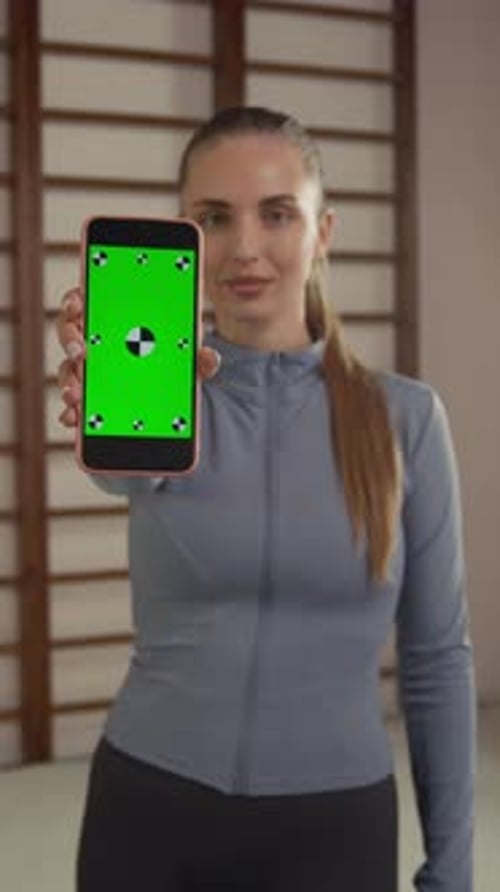 Young Fit Woman Demonstrating Smartphone with Green Screen In Sports Studio