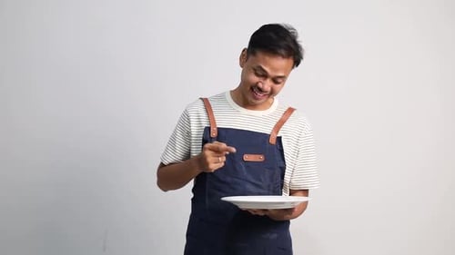 A young male restaurant waiter carries an empty plate while pointing at the plate he is carrying.