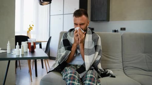 Sick Man Sneezing Into Tissue Covered in Blanket on Couch