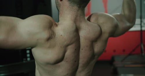 Strong and muscular shirtless bodybuilder is pulling on weight machine inside gym to train his back