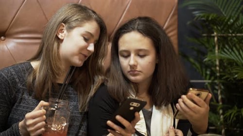 Teenage girlfriends chatting over smartphones and juice in a sunny cafe