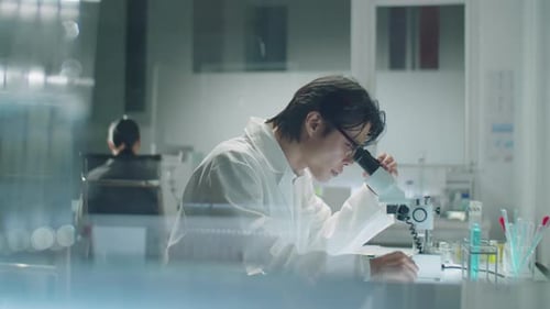 Asian Scientist Using Microscope and Making Notes in Laboratory