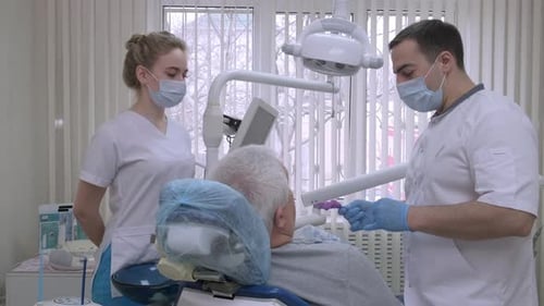 Dentist with Nurse in Dental Clinic Explaining Artificial Teeth to Senior Man