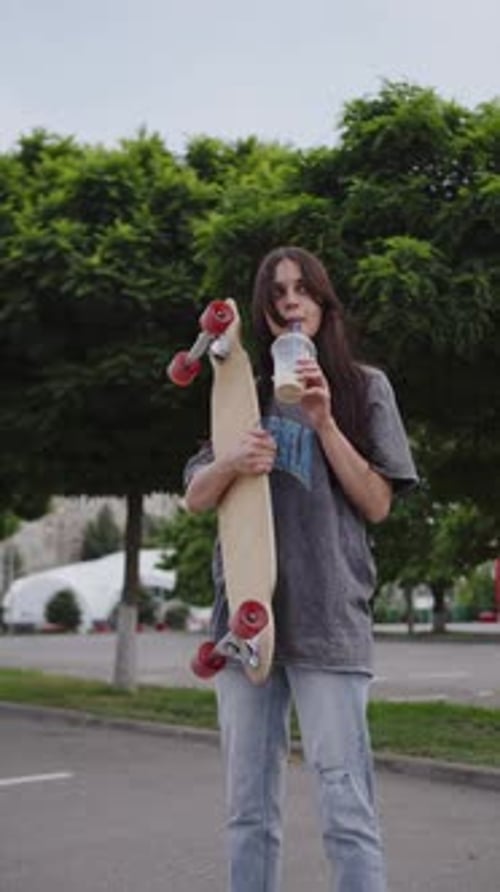 Young Girl with a Longboard and Coffee in Hand Stands in the Parking Lot Vertical Video