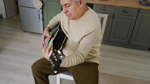 Senior Man Sitting Playing Acoustic Guitar in Kitchen