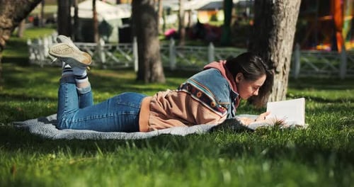 Portrait of Cheerful Young Woman Reading Book Laying on the Grass Reading Outside in Spring Sunlight
