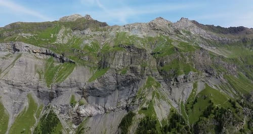 Aerial shot showcasing a dramatic alpine cliff with exposed geological layers and steep grassy