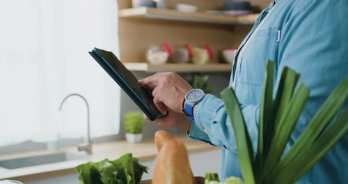 Close up unrecognizable man tapping on tablet device browsing online standing in modern kitchen.