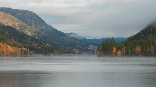 Forest-covered mountains and clouded sky reflected in the still mirrorlike waters of the lake. Thin