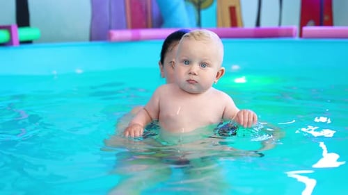 Beautiful blond toddler is in the swimming pool. Female coach is holding is lifting a child up.
