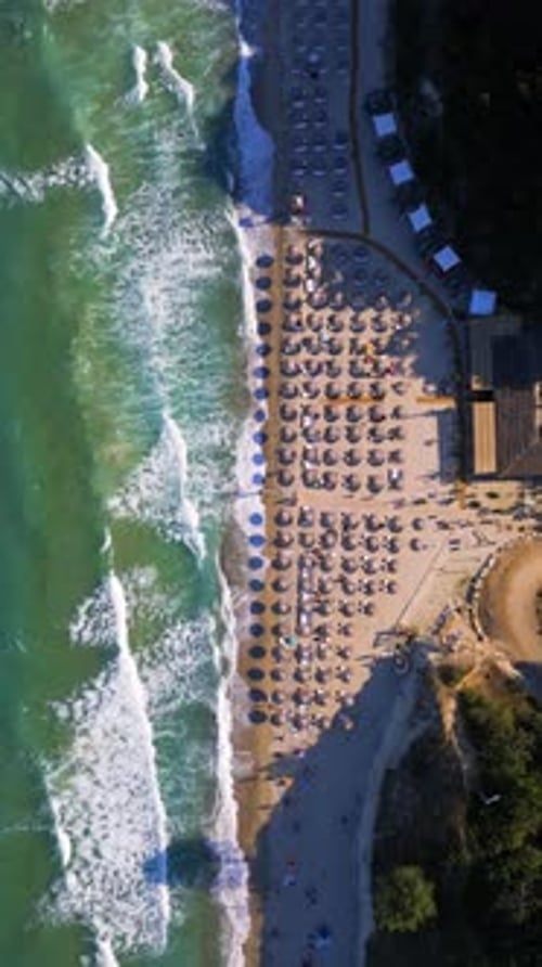 Top-down aerial video of a busy beach with rows of umbrellas and sunbeds, waves crashing along the s