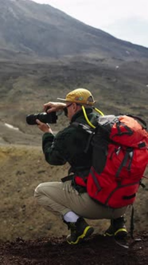Photographe en plein air avec sac à dos de randonnée rouge accroupi sur le versant d'une montagne rocheuse capturant des images théâtrales