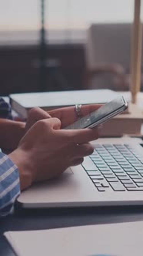 Close Up of Hands Using a Smartphone Over a Laptop Keyboard in a Modern Office Workspace