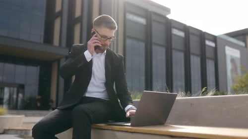 Professional Man in Suit Using Laptop and Phone on Bench in Busy City