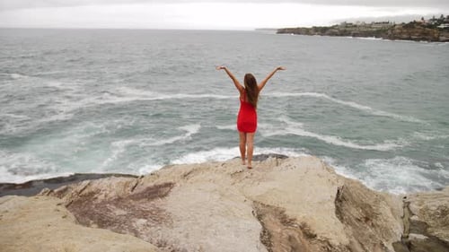 Sexy Girl In Red Dress Stands On The Edge Of A Rocky Coast And Raised Both Hands - Ocean Waves Cras