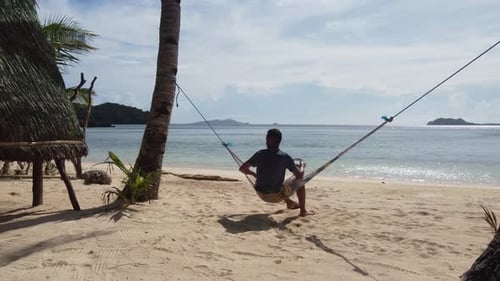 Young adult swing on tree hammock at tropical sunny sand beach beside bungalow