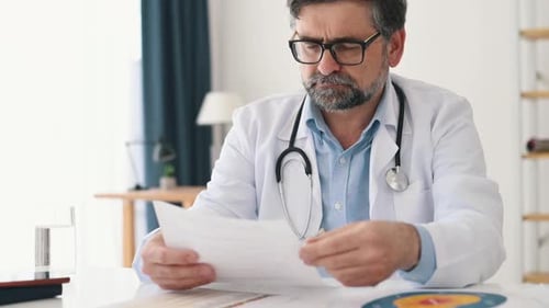 Reading the document. Mature male doctor in coat is in the clinic room