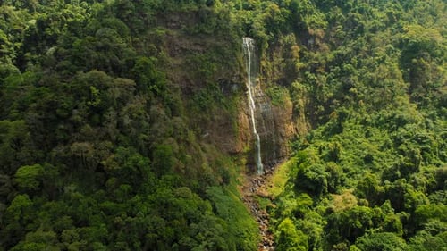 Drone showing waterfall flowing through steep rocky cliffs and dense lush forest