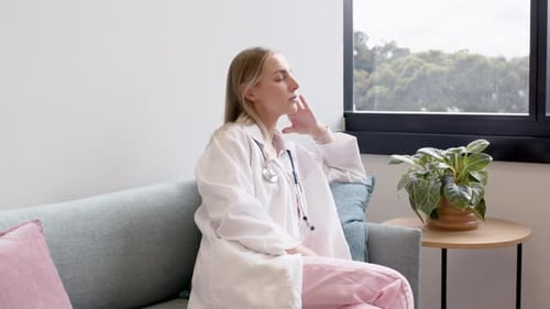 Erwachsene Ärztin sitzt auf dem Kliniksofa und blickt mit Stethoskop in Richtung Baumfenster