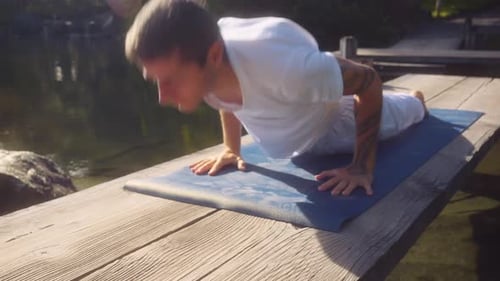 Caucasian adult man does yoga and stretches on wooden bridge over small pond in lush garden park. Su