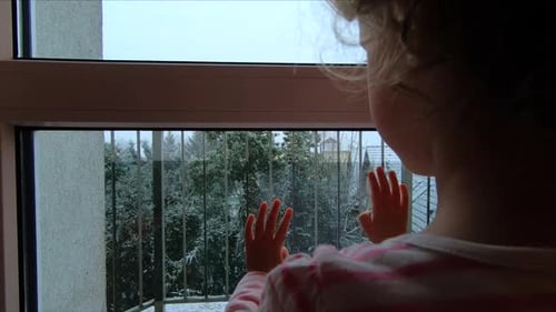 A little girl is watching through the window the snow falling