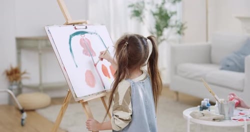 Creative playtime in living room as preschool daughter paints shape on easel canvas, Asian mother