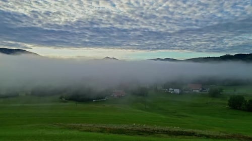 Misty valley at sunrise with clouds overhead and scattered houses, aerial view