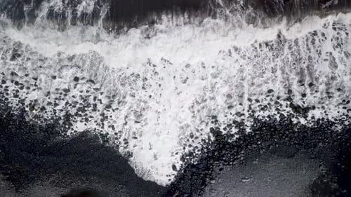 Monochrome top-down view of powerful foamy waves hitting a rocky black sand beach in Maui, Hawaii