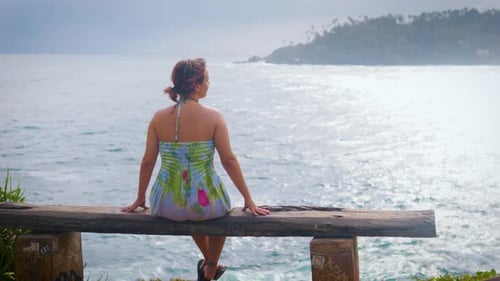 Mädchen sitzt auf einer Bank an der Küste Siziliens, Italien, blickt auf das Meer und den Ozean von hinten