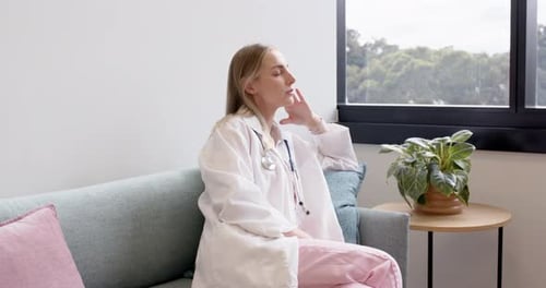 Erwachsene Ärztin sitzt auf dem Kliniksofa und blickt mit Stethoskop in Richtung Baumfenster