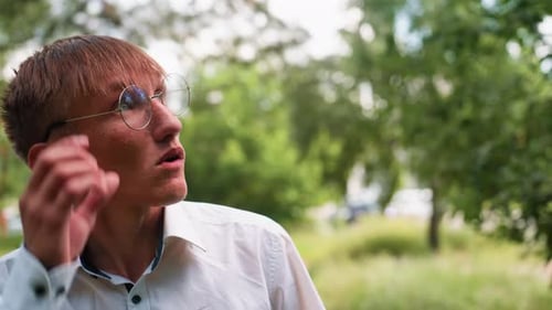 Portrait of Young Man in Glasses Observing Trees with Blurred Cars Behind
