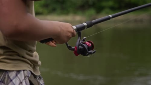 Hands of Teenage Boy Winding Reel on Angling Rod while Fishing on River